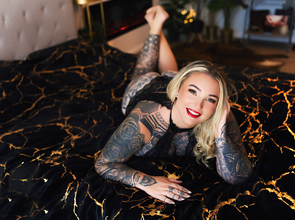 Boudoir client with tattoos resting on bed, smiling. Fun photo shoot experience.